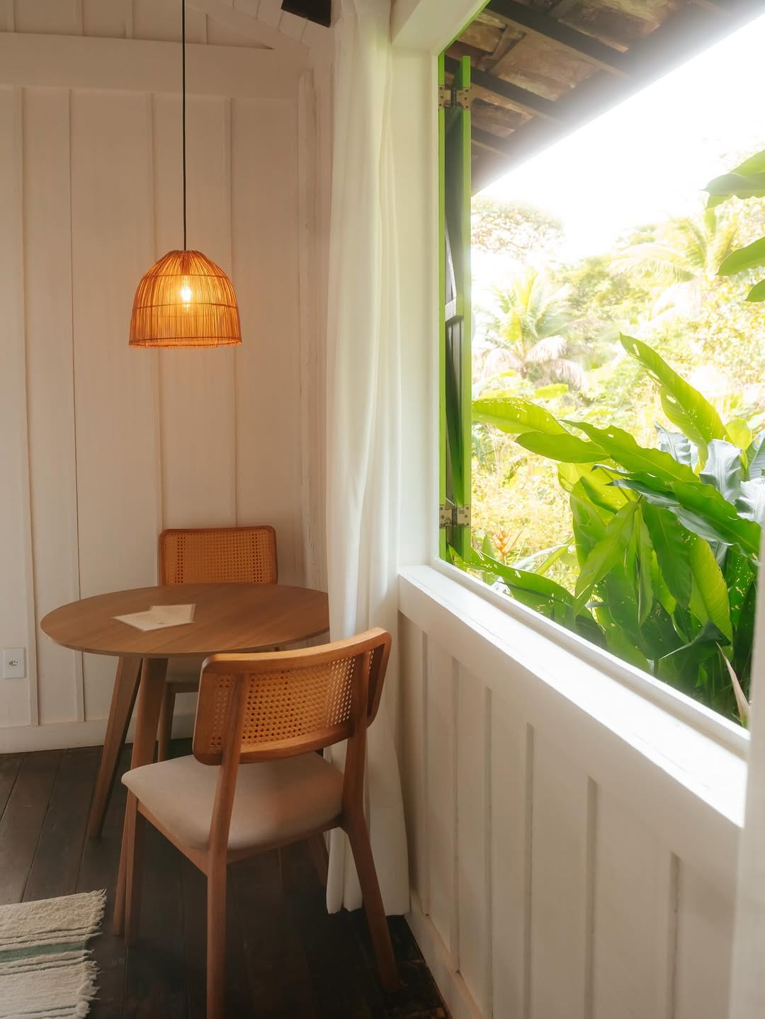 Mesa de madeira com cadeiras e luminaria artesanal junto a janela com vista para a mata